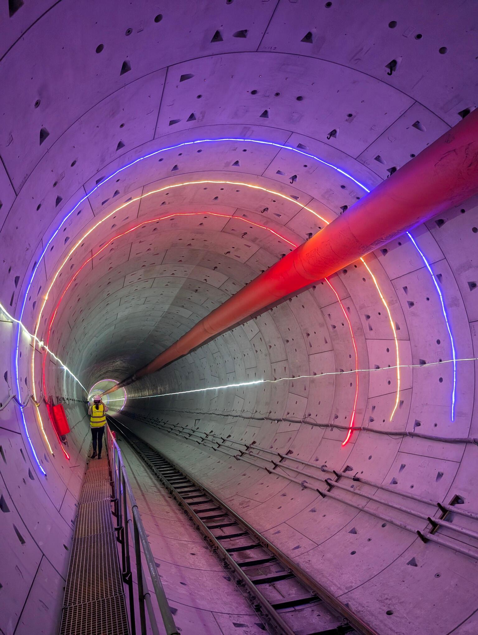 A perspective view looking down a finished concrete railway tunnel, illuminated by colorful neon ring lights (blue, orange, and red) and featuring a worker walking along a side maintenance platform