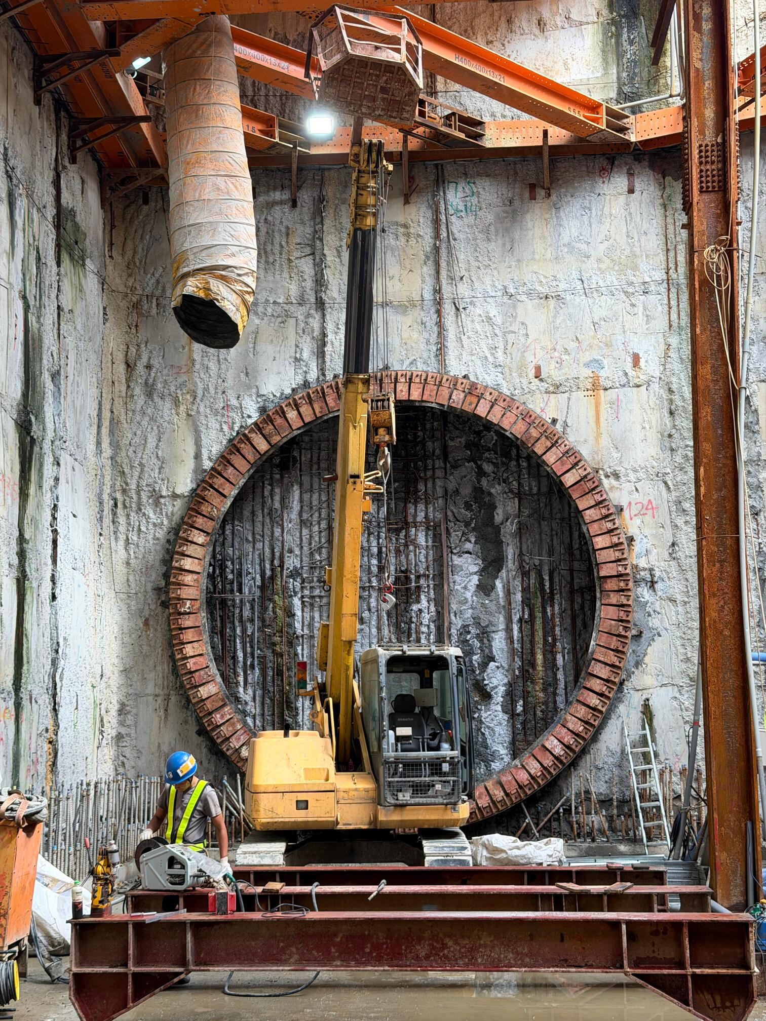 An excavator stands in front of a circular opening in a concrete wall at an underground construction site
